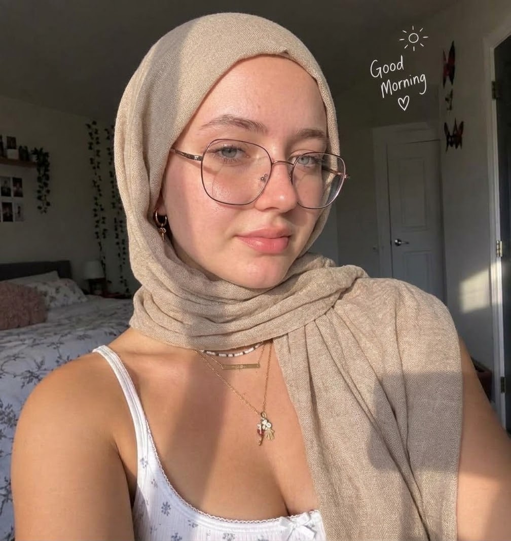 Sunlit bedroom selfie with a beige hijab, round glasses, pale floral top and layered necklaces.