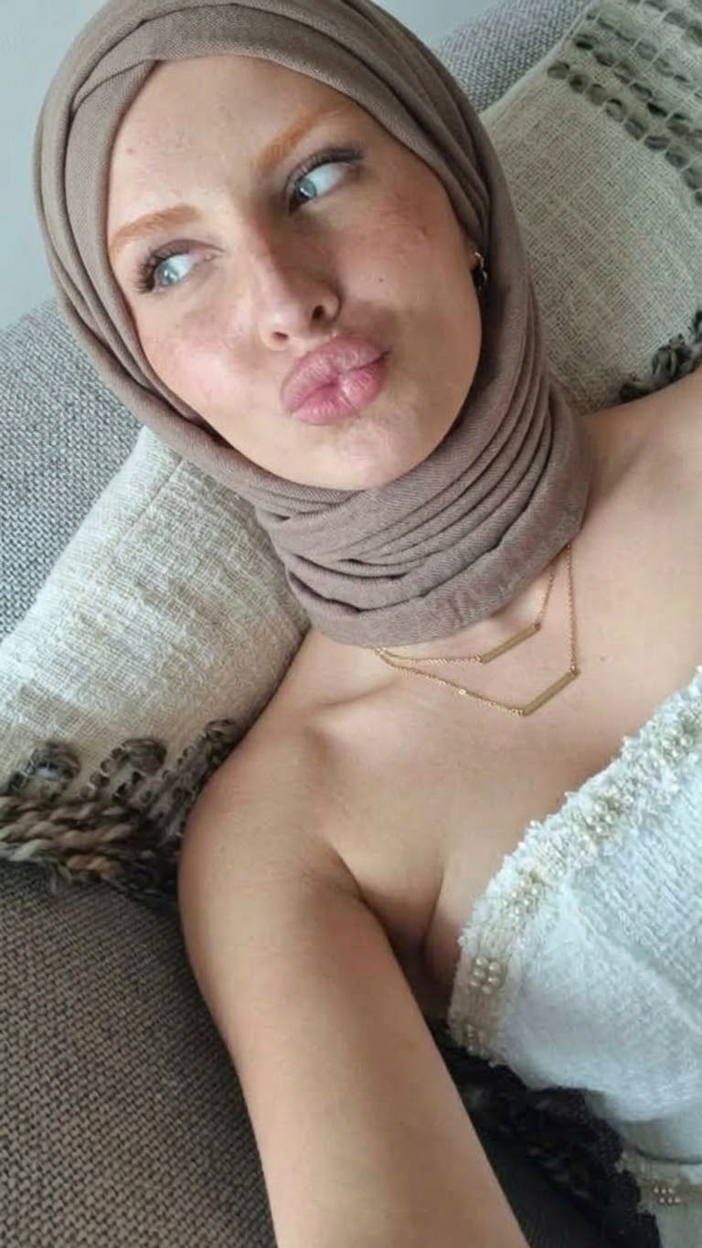 Reclining sofa selfie with a taupe hijab, fluffy white outer layer and puckered lips beside textured cushions.