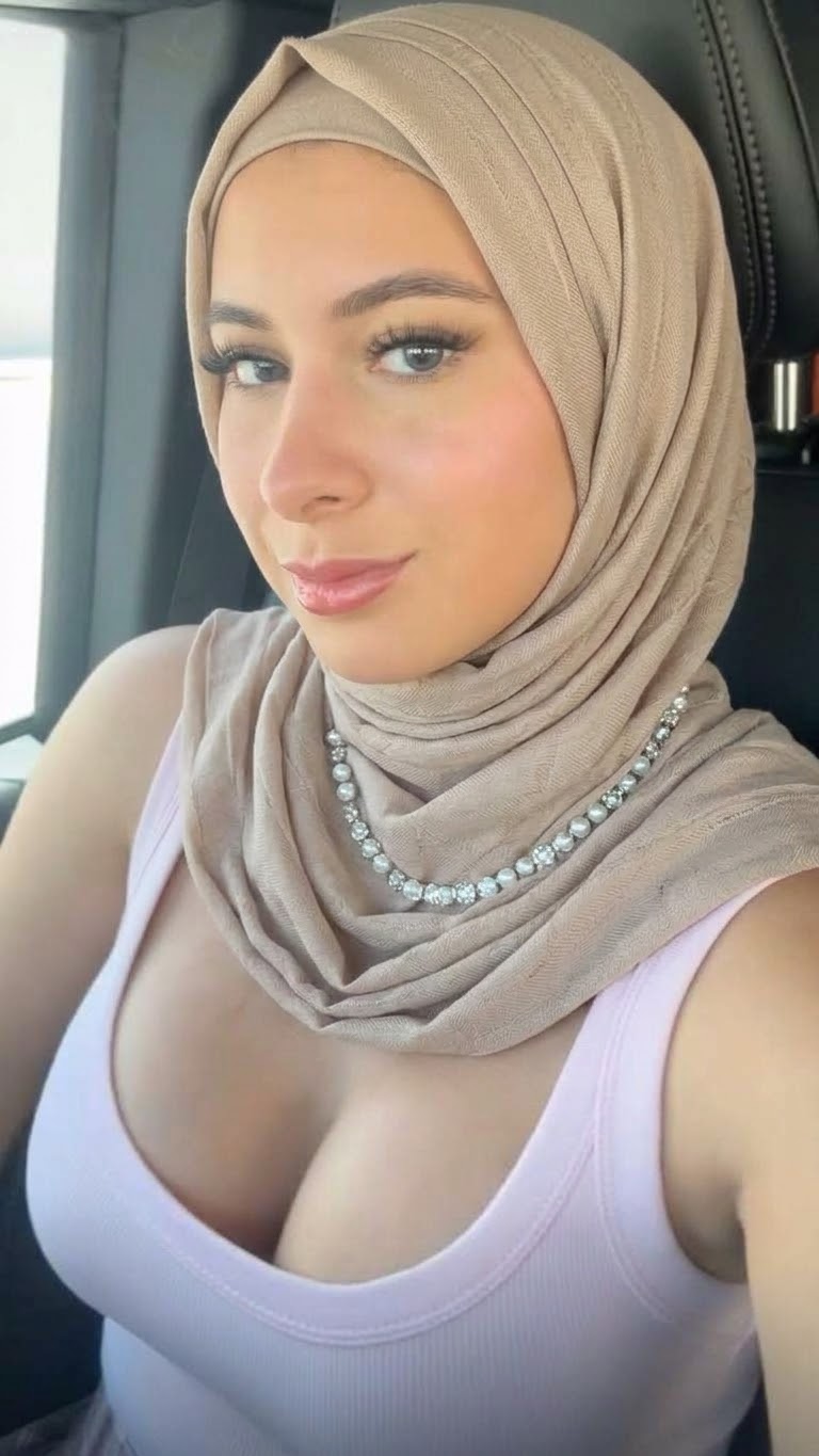 Window-lit selfie with a beige hijab, pale scoop-neck tank top and layered necklaces, looking sideways.