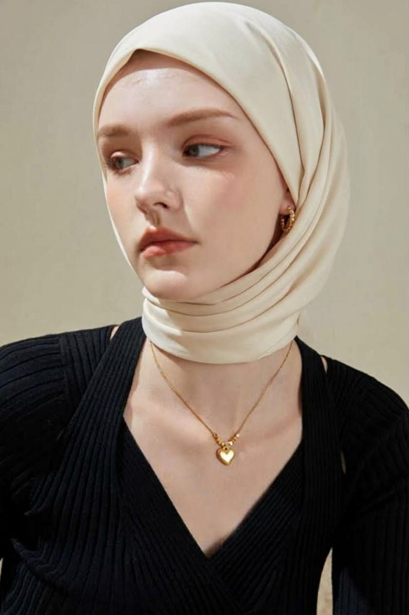 حجاب كريمي مع بلوزة سوداء مضلعة بياقة V وقلادة قلب ذهبية ونظرة جانبية.