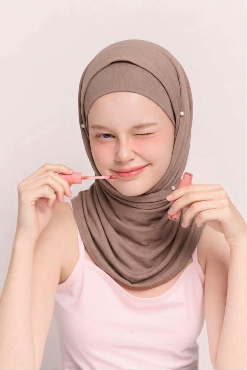 بورتريه بغمزة مع حجاب بلون تاوب وبلوزة وردية فاتحة بحمالات ونظارة تمسكها اليدان.