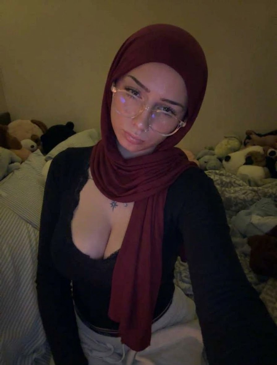 Burgundy hijab, wire-frame glasses and black long-sleeve V-neck top in a tilted indoor portrait.