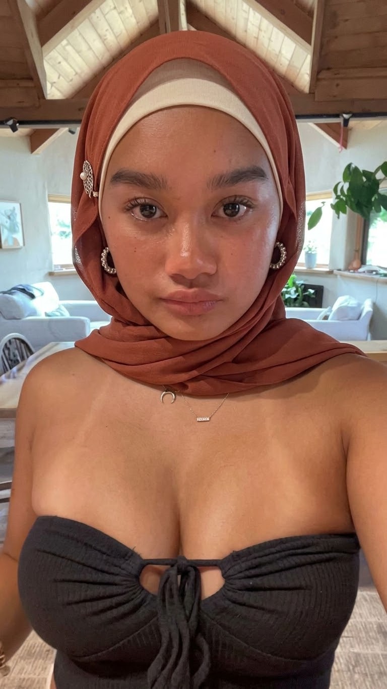 Rust-burgundy hijab with a white undercap, hoop earrings and knotted black strapless top beneath timber beams.