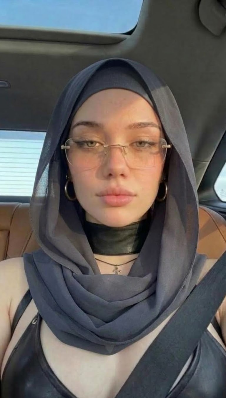 سيلفي أمامي داخل السيارة بحجاب رمادي أردوازي ونظارة معدنية وحزام أمان وسماء زرقاء عبر فتحة السقف.