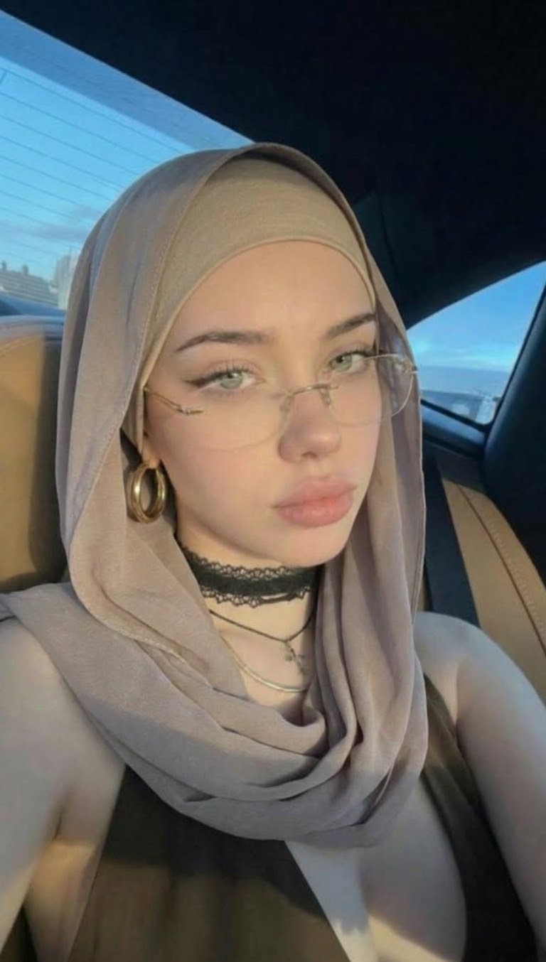 سيلفي مائل داخل السيارة مع حجاب رمادي مائل للبيج ونظارة وأقراط ذهبية وقلادة دانتيل وحزام أمان.