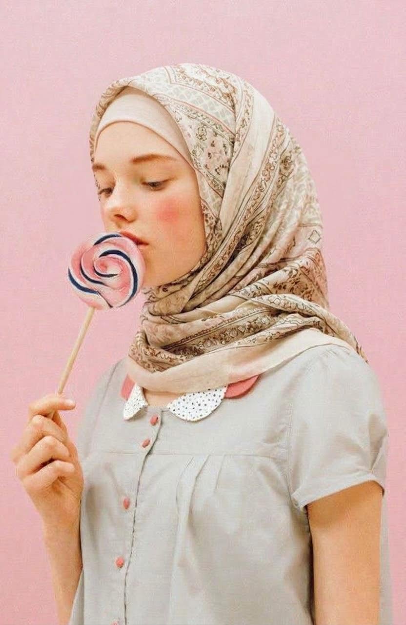 Patterned beige hijab and gray blouse with closed eyes and a swirl lollipop against pink.